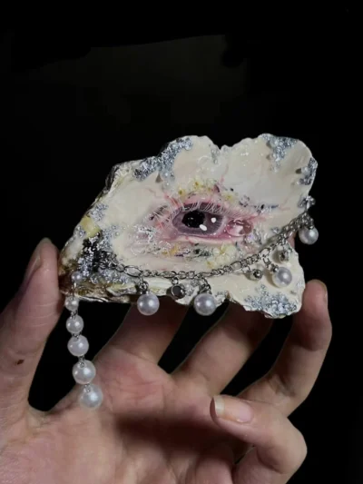 An artist's hand holding 'The Gilded Gaze' oyster shell eyeball sculpture, demonstrating its scale and pearl dangles. A rare post-apocalyptic curiosity.