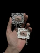 An artist's hand holding two of the seashell eyeball sculptures, showcasing the scale, intricate baroque textures, and the delicate pearl and chain embellishments. Vulture culture art alternative.