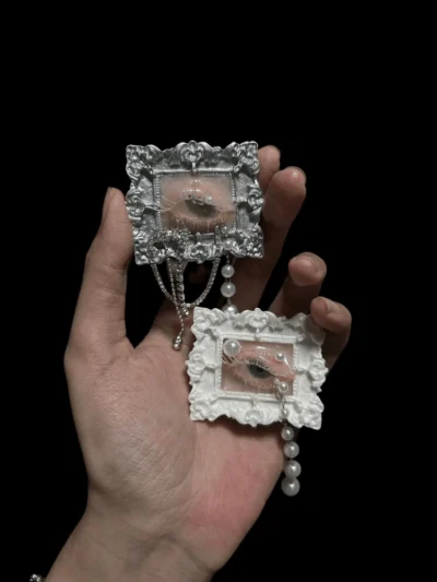 An artist's hand holding two of the seashell eyeball sculptures, showcasing the scale, intricate baroque textures, and the delicate pearl and chain embellishments. Vulture culture art alternative.