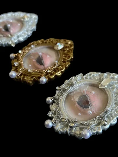Three handcrafted miniature realistic human seashells eyes, meticulously sculpted, encased in ornate Baroque-style frames: one gold, two silver, on a black background.