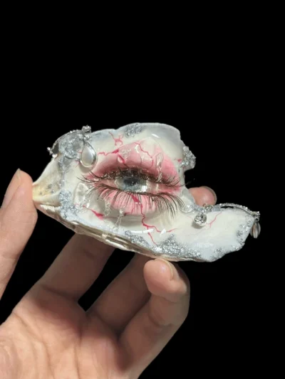Handcrafted seashell sculpture featuring a hyper-realistic crying human eye with long lashes, red veins, silver glitter accents, and a pearl dangle, held by a hand. A unique Vulture Culture oddity.