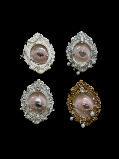 Collection of four handcrafted realistic seashell eyeball miniatures in ornate frames: white with crosses, silver with crystals, silver with pearls, and gold with pearls, against a black background.