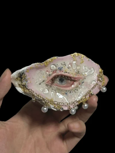 A macro photograph of a realistic human eye sculpture with detailed eyelashes, set inside a pink-and-gold gilded seashell, featuring a crystal floral 'nest' and dangling pearl charms, held by a hand. A unique piece of whimsigoth or dark academia room decor.