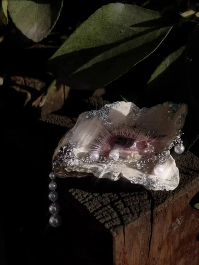 Side profile of 'The Gilded Gaze' shell art on a dark wood block under a patch of dappled sunlight. A perfect creepy-cute addition to a gothic room.