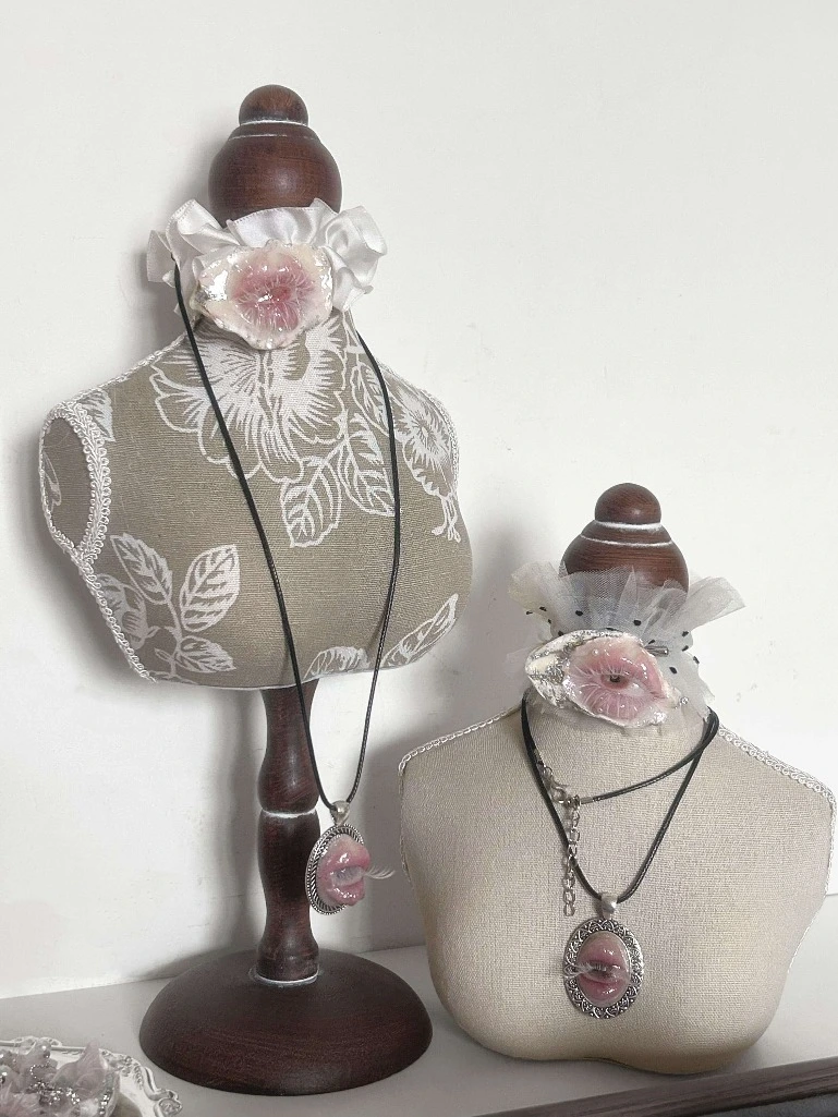 Bizarre and surreal art jewelry display. Miniature mannequins wearing creepy-cute fleshy pink lip pendants, styled with dark cords and ruffled white tulle for an alternative, gothic-inspired look.