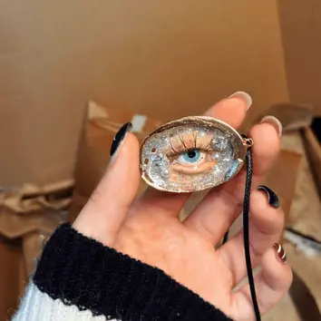 Hand holding a gothic handmade shell necklace with a sculpted blue eye, silver glitter, and pearl accents on a black cord.