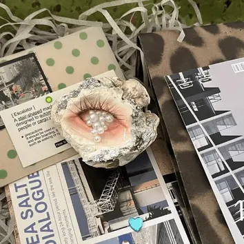 Creepy cute handmade oyster shell sculpture of a pink eye crying pearls, resting on modern aesthetic magazines and postcards.