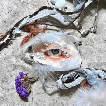 Quirky handmade shell pendant with a realistic hazel eye, decorated with white feathers and pearls, displayed with a dried purple flower.