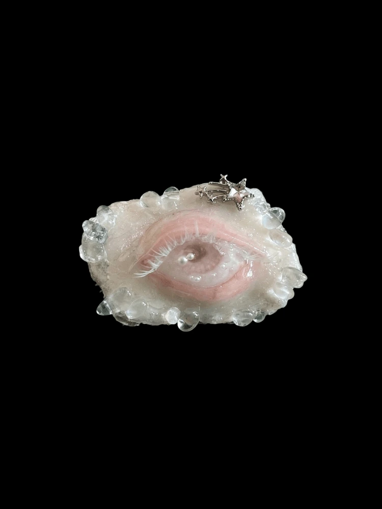 Close-up of a hand-sculpted, weeping Lover's Eye brooch with resin tears and a star charm, against a black background.