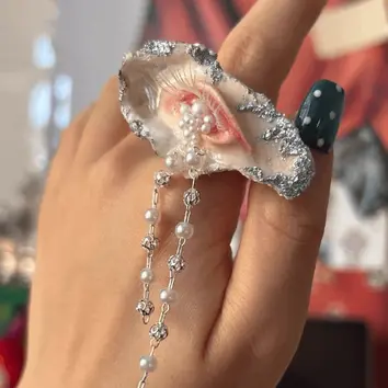 Handmade surreal oyster shell ring featuring a sculpted realistic eye crying pearl tears with silver glitter edges, worn on a hand.