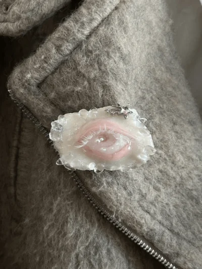 A high-fashion, weirdcore Lover's Eye brooch worn on the lapel of a gray wool coat, styled as an oddity accessory.