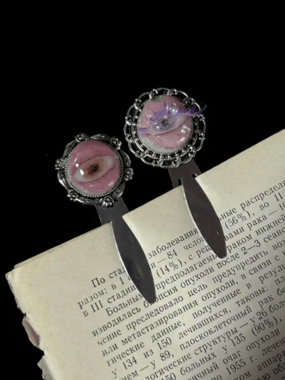 Handcrafted seashell eyeball bookmarks with detailed eyelashes and pearl adornments resting among aged, text-heavy antique book pages.